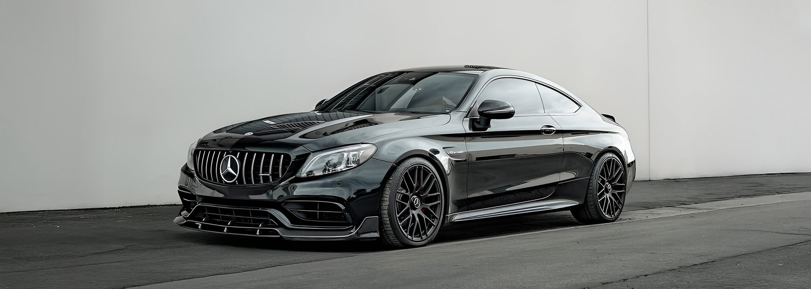 Mercedes C63 Coupe with Carbon Noire carbon fibre front splitter, side skirts, and rear spoiler parked against a minimalist wall